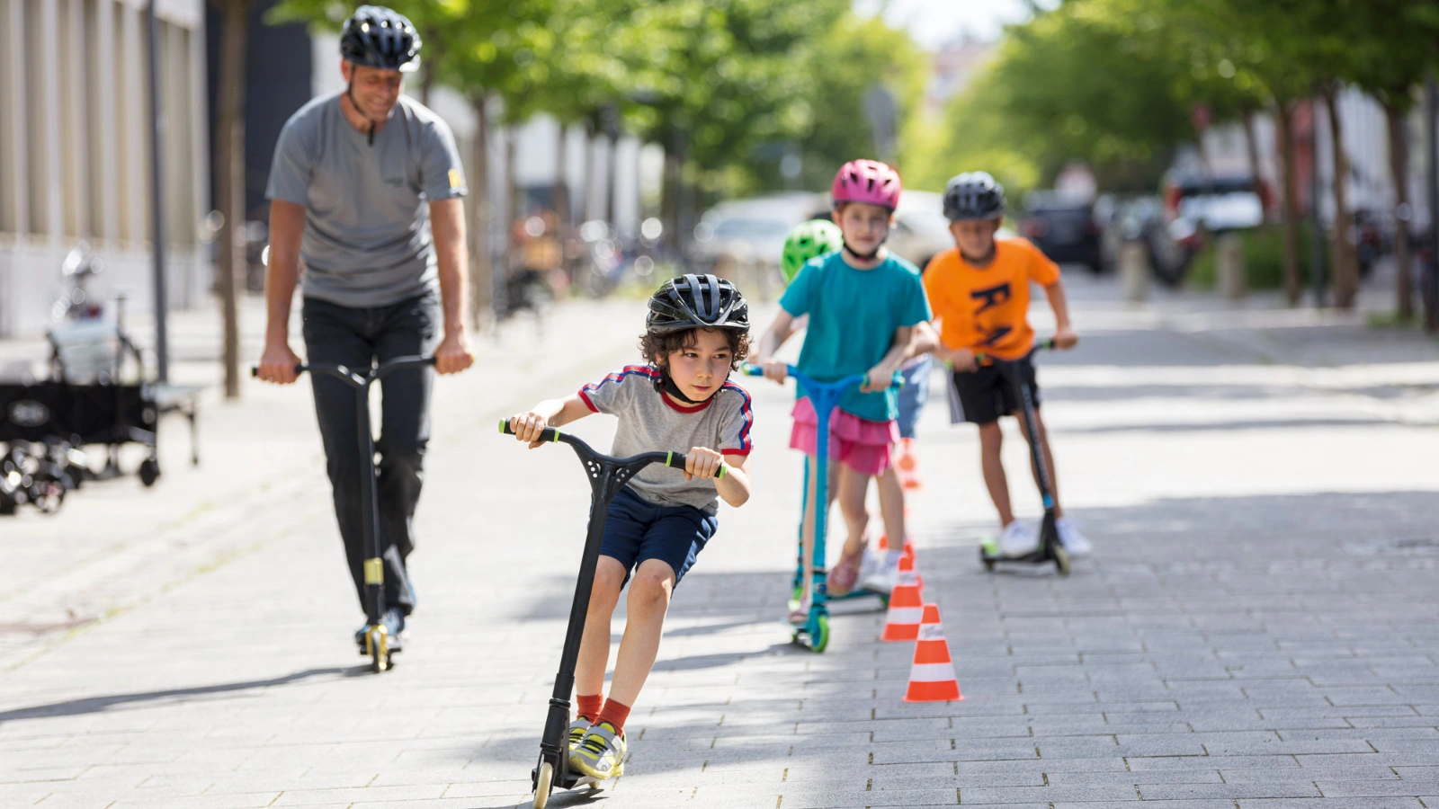 Bild zeigt Kinder mit Helm beim Rollerfahren um Pylonen. Eine Lehrkraft begleitet sie.