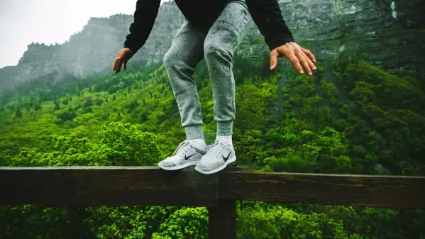 Bild zeigt eine Person, die auf einem Brückengeländer balanciert. Im Hintergrund sind Berge und Wälder zu sehen.